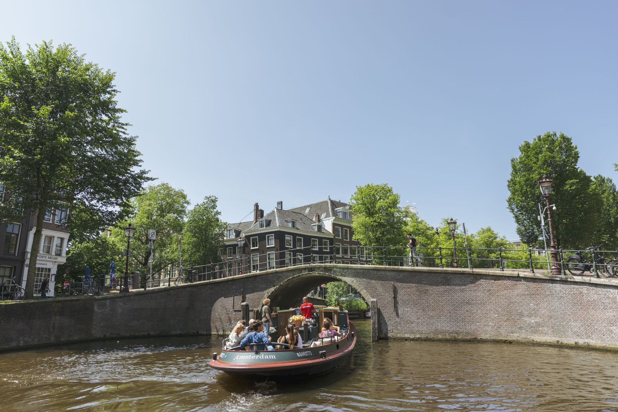 Boaty.nl - Rondvaart boot op gracht A boat full of passengers sails under a stone bridge on the Amsterdam canals, surrounded by historic buildings and greenery.