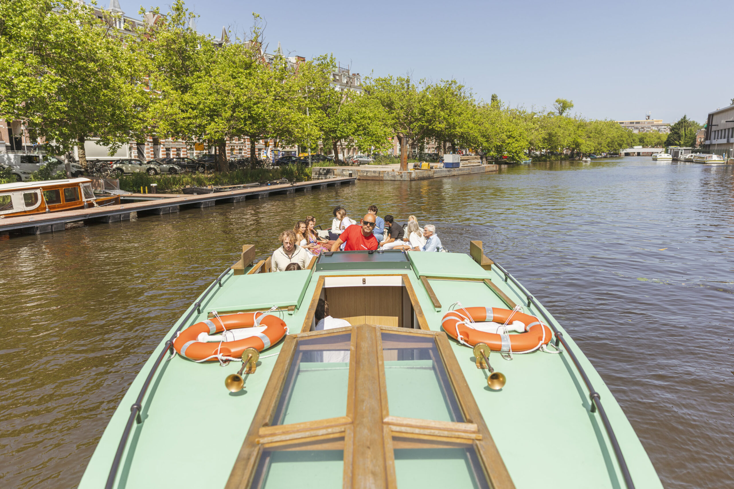 Boaty.nl - Rondvaartboot met schipper A group of people sit relaxed on a classic canal boat in Amsterdam, as the skipper steers the vessel through the canal on a sunny day.