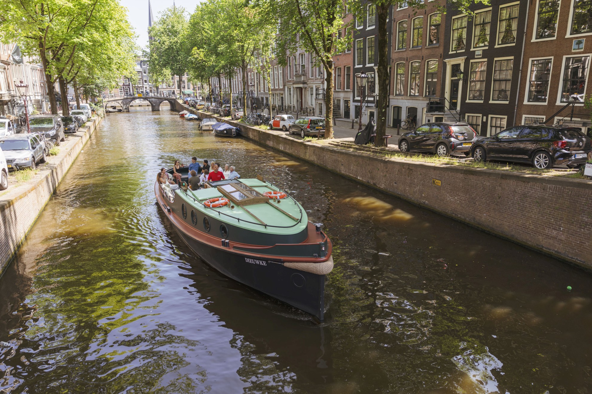 Boaty.nl - Rondvaartboot op Amsterdamse gracht A group of people sails through an Amsterdam canal in a classic boat, surrounded by trees, parked cars, and historic canal houses.
