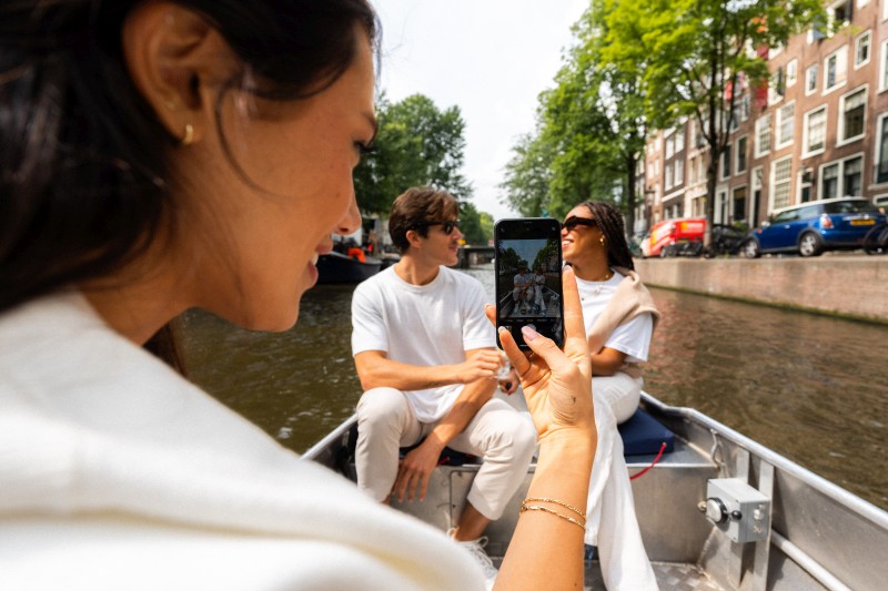 Friends take a photo during a relaxed boat trip through an Amsterdam canal, surrounded by historic canal houses and green trees.