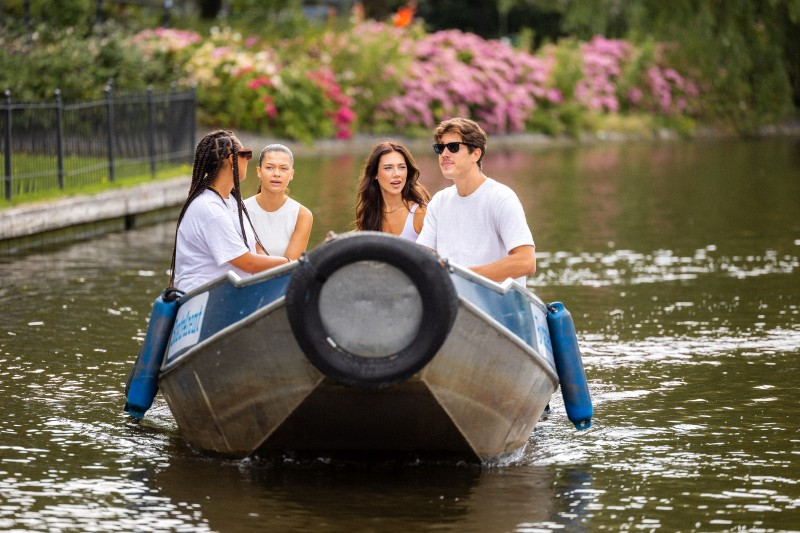 Four friends are sailing together in a small boat along a quiet canal, surrounded by greenery and flowering bushes.