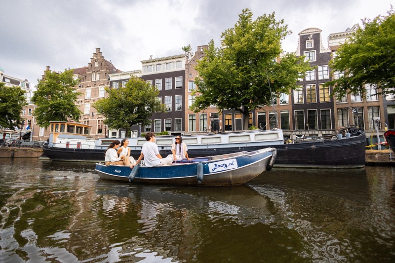 A group of friends sails in a small rental boat through an Amsterdam canal, past historic canal houses and houseboats.