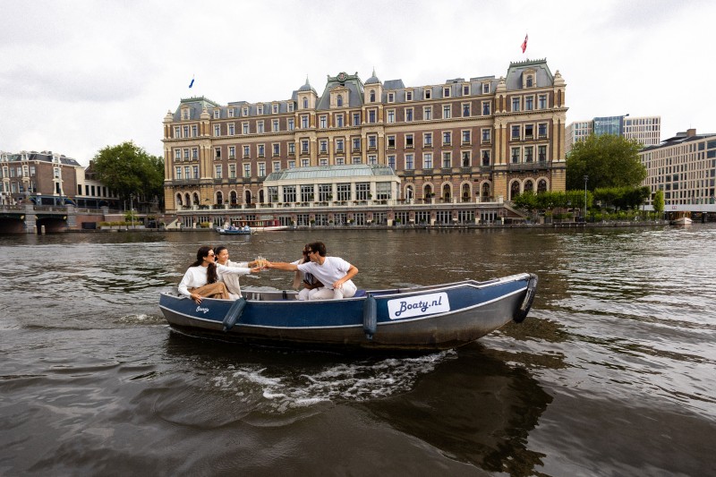 Friends toast with drinks during a boat trip on the water with a historic building along the Amsterdam canal in the background.