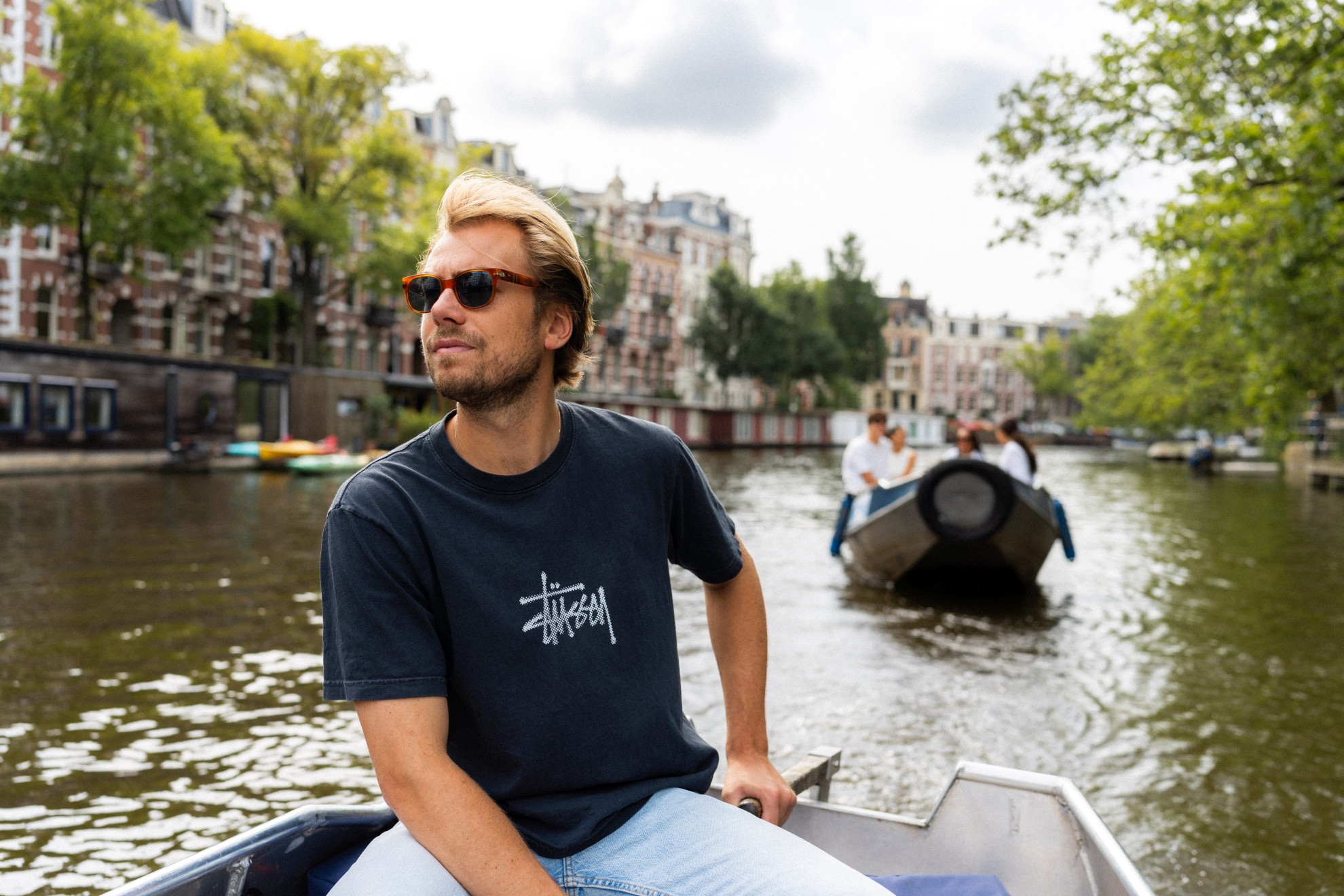 A man is steering a boat on the Amsterdam canals, and another small boat is following him on the water.